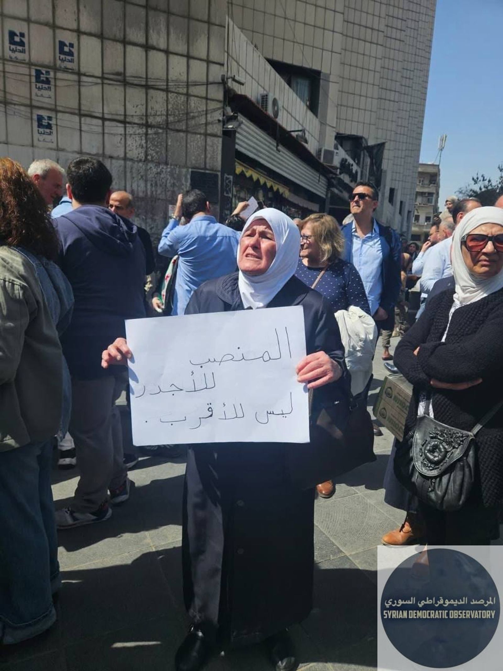 سيدة مسنة في تحمل لوحة كتب عليها "المنصب للأجدر وليس للأقرب"