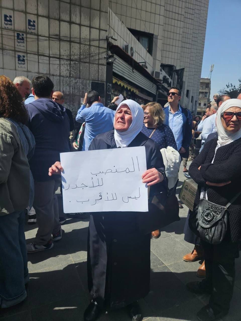 سيدة مسنة في تحمل لوحة كتب عليها "المنصب للأجدر وليس للأقرب"