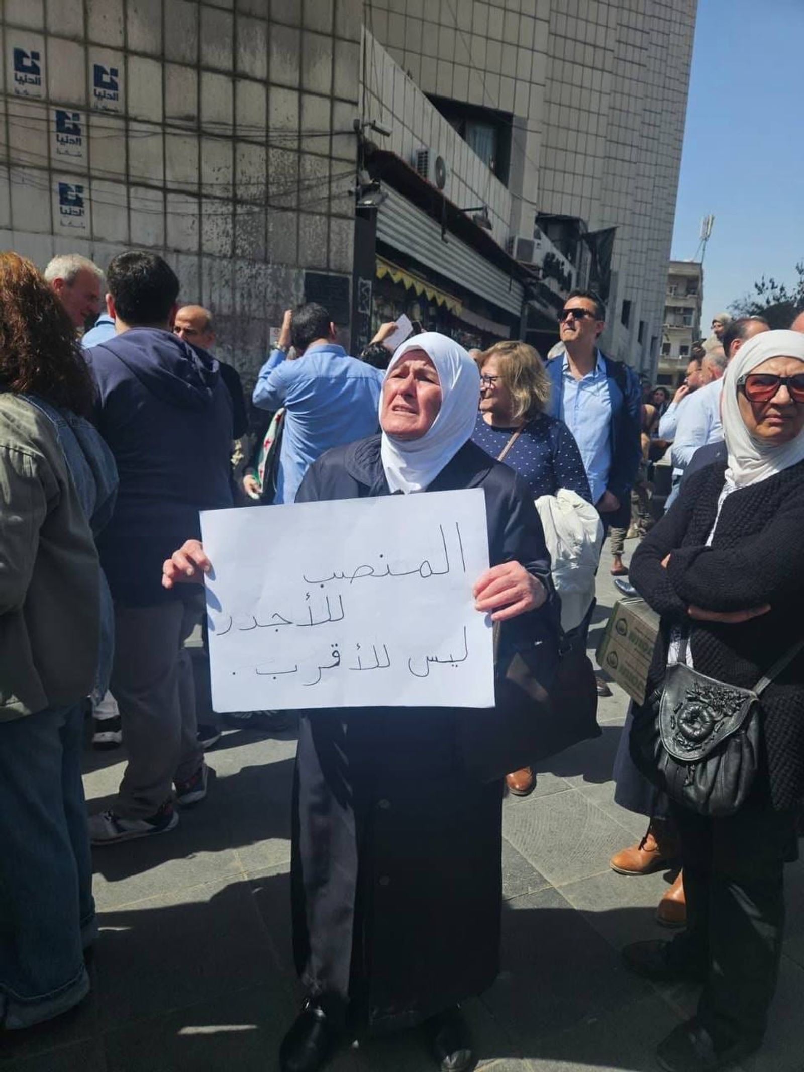 سيدة مسنة في تحمل لوحة كتب عليها "المنصب للأجدر وليس للأقرب"